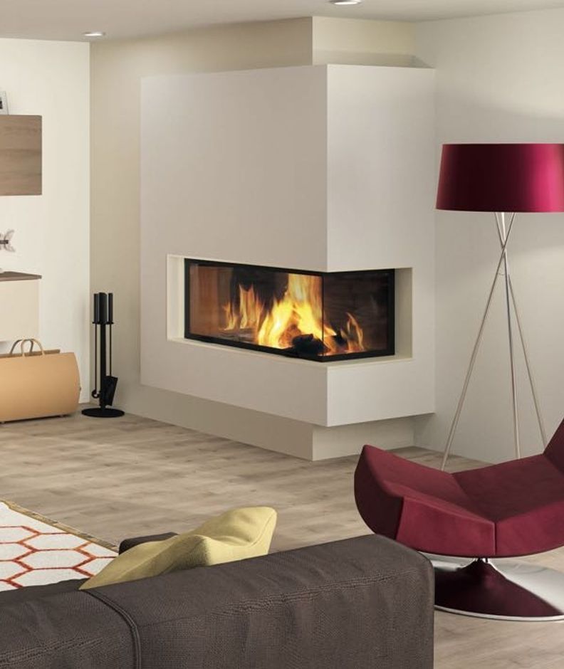Cheminée d'angle moderne avec flammes, encastrée dans un mur blanc. Fauteuil et lampe bordeaux, parquet clair.