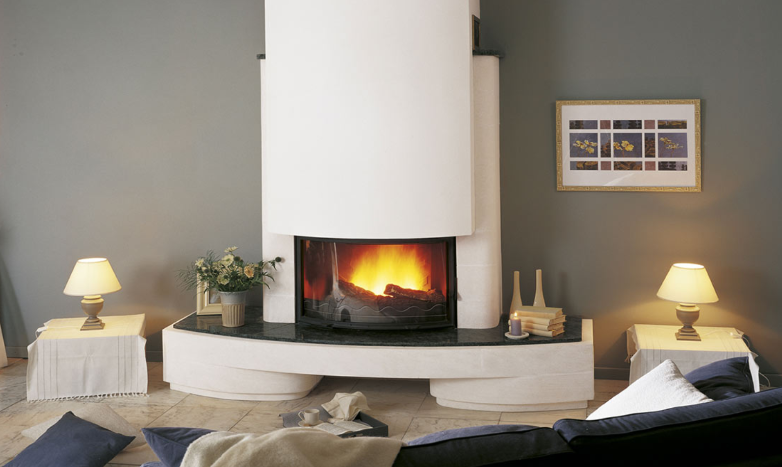 Cheminée blanche avec un feu, flanquée de lampes sur de petites tables, contre un mur gris.