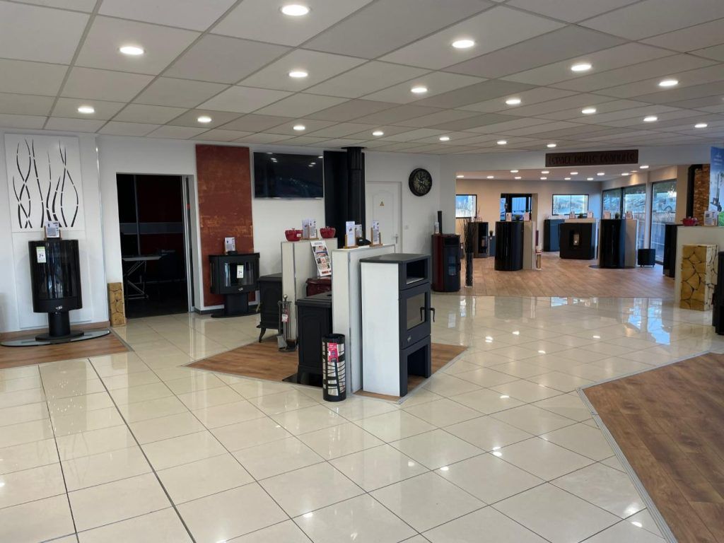 Intérieur d'une salle d'exposition présentant divers poêles à bois sur un sol carrelé sous un éclairage vif au plafond.