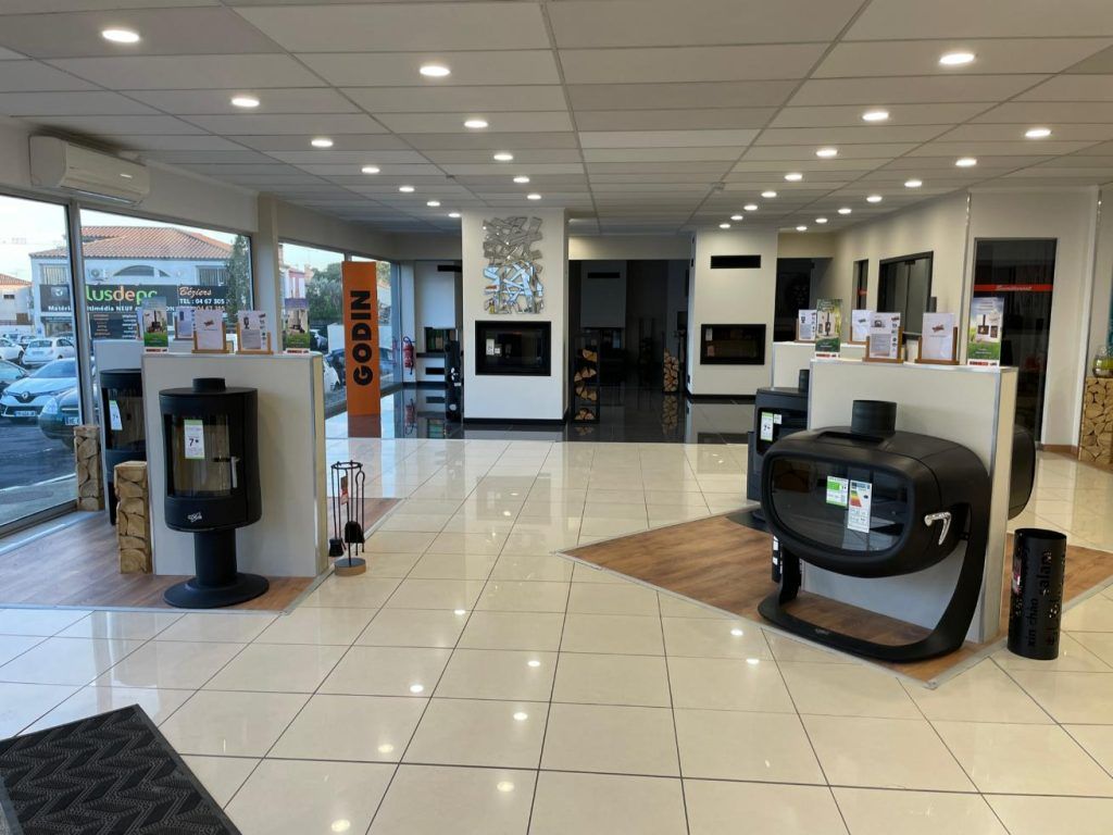 Intérieur de la salle d'exposition avec poêles à bois et cheminées exposés, lumineux grâce à de grandes fenêtres.