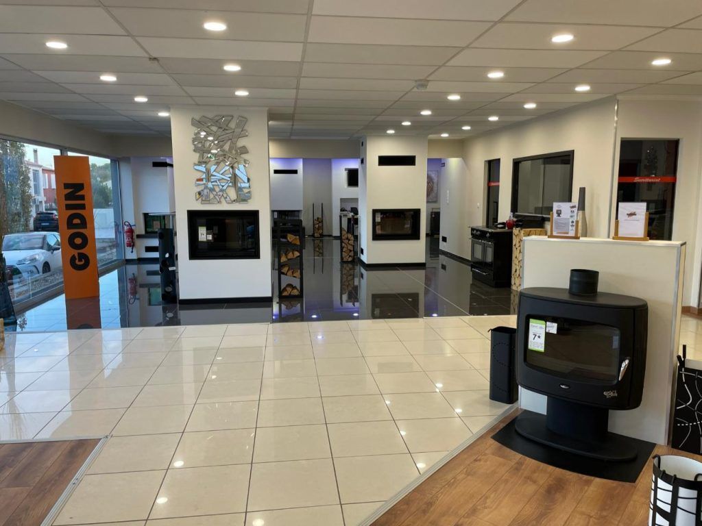 Intérieur d'une salle d'exposition présentant divers foyers et poêles ; lumineux grâce à son sol réfléchissant. Enseigne GODIN à gauche.