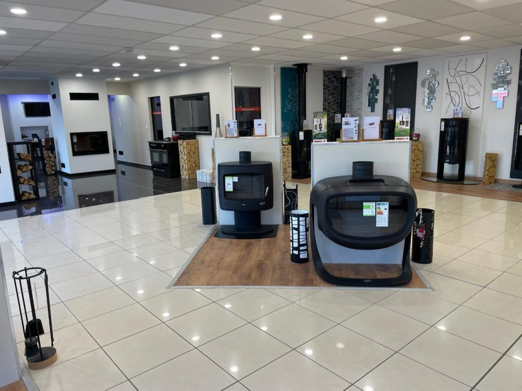 Salle d'exposition présentant plusieurs poêles à bois et cheminées, avec sol carrelé blanc et éclairage zénithal lumineux.