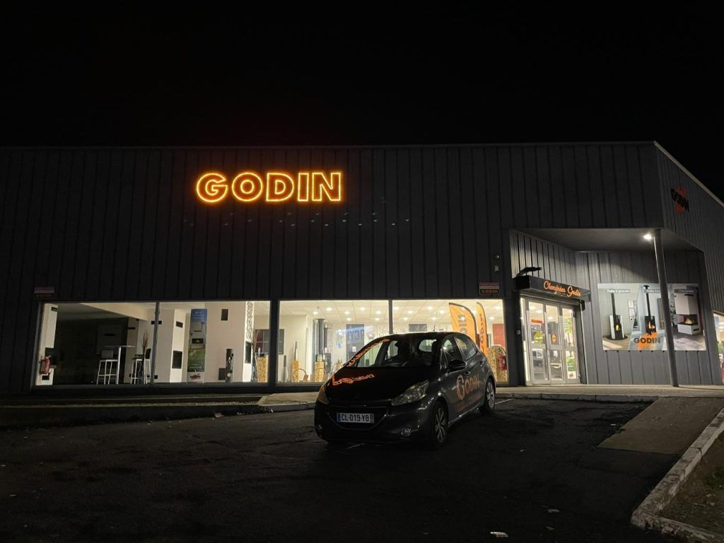 Bâtiment avec une enseigne lumineuse Godin, voiture sombre garée devant. La nuit.