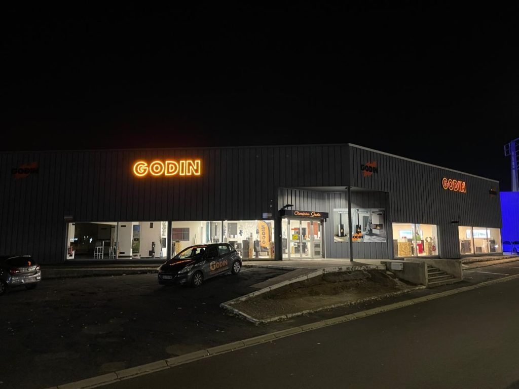Vue nocturne du magasin Godin, avec son enseigne et son entrée illuminées. Des voitures sont garées devant.