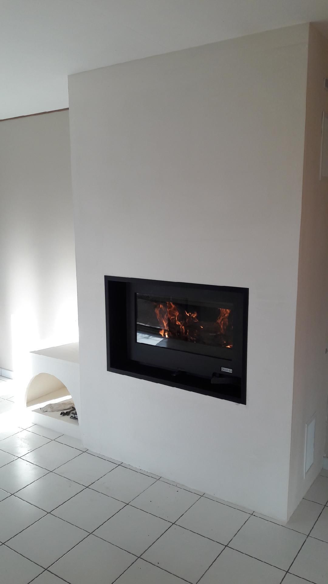 Cheminée encastrée dans un mur blanc, intérieur noir. Les flammes sont visibles. Sol carrelé blanc.