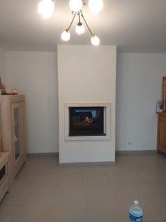 Salon avec cheminée, murs blancs et sol carrelé. Un feu brûle dans la cheminée.