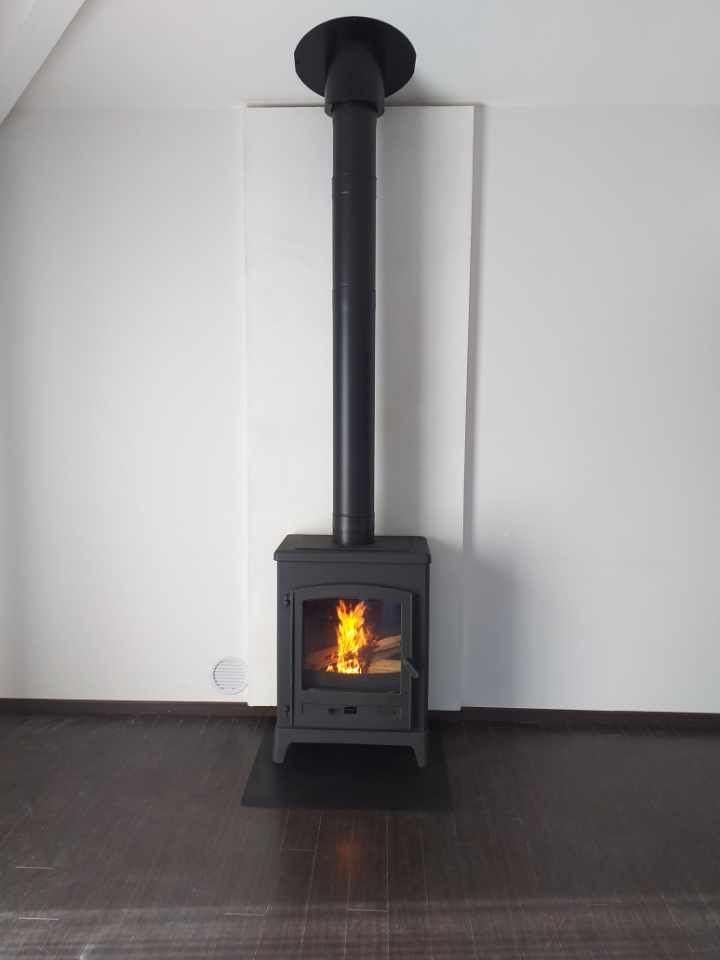 Poêle à bois avec flammes, cheminée noire, sur sol sombre, contre un mur blanc.