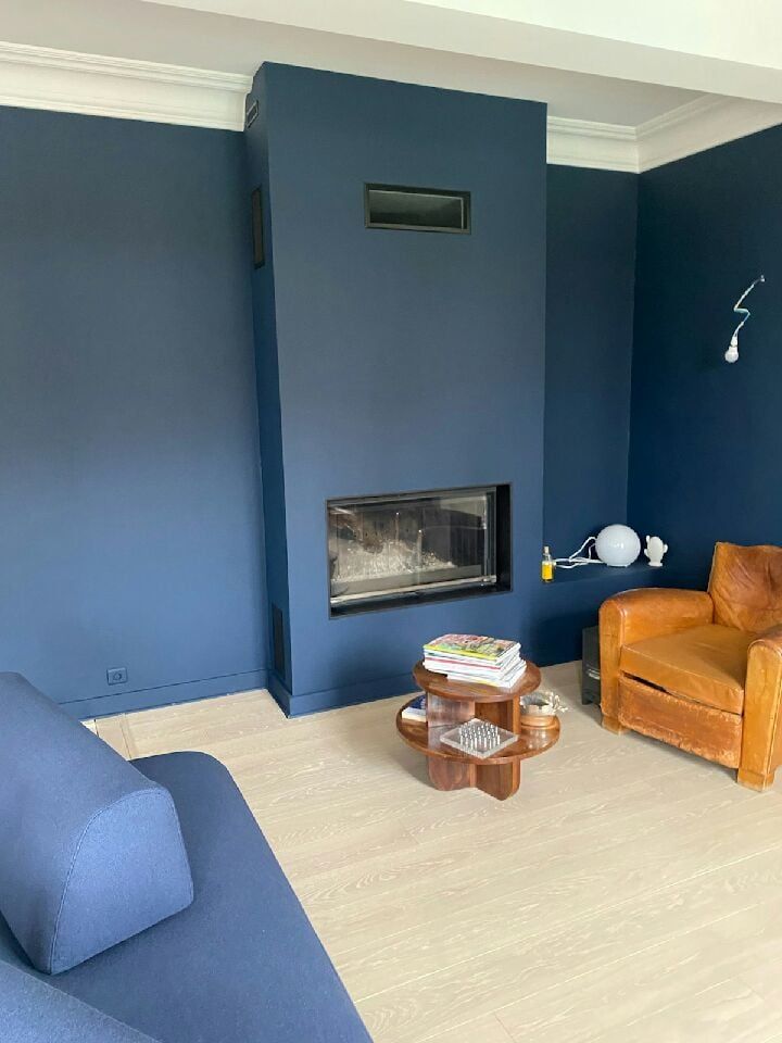 Salon aux murs bleus avec cheminée moderne, fauteuil en cuir, table basse en bois et sol clair.