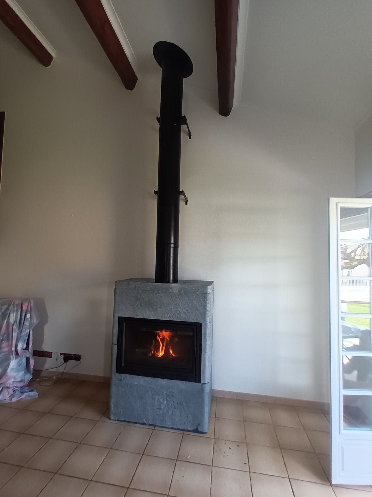 Poêle à bois avec feu allumé, cheminée noire, façade en pierre grise. Sol carrelé beige et murs blancs, poutres apparentes.