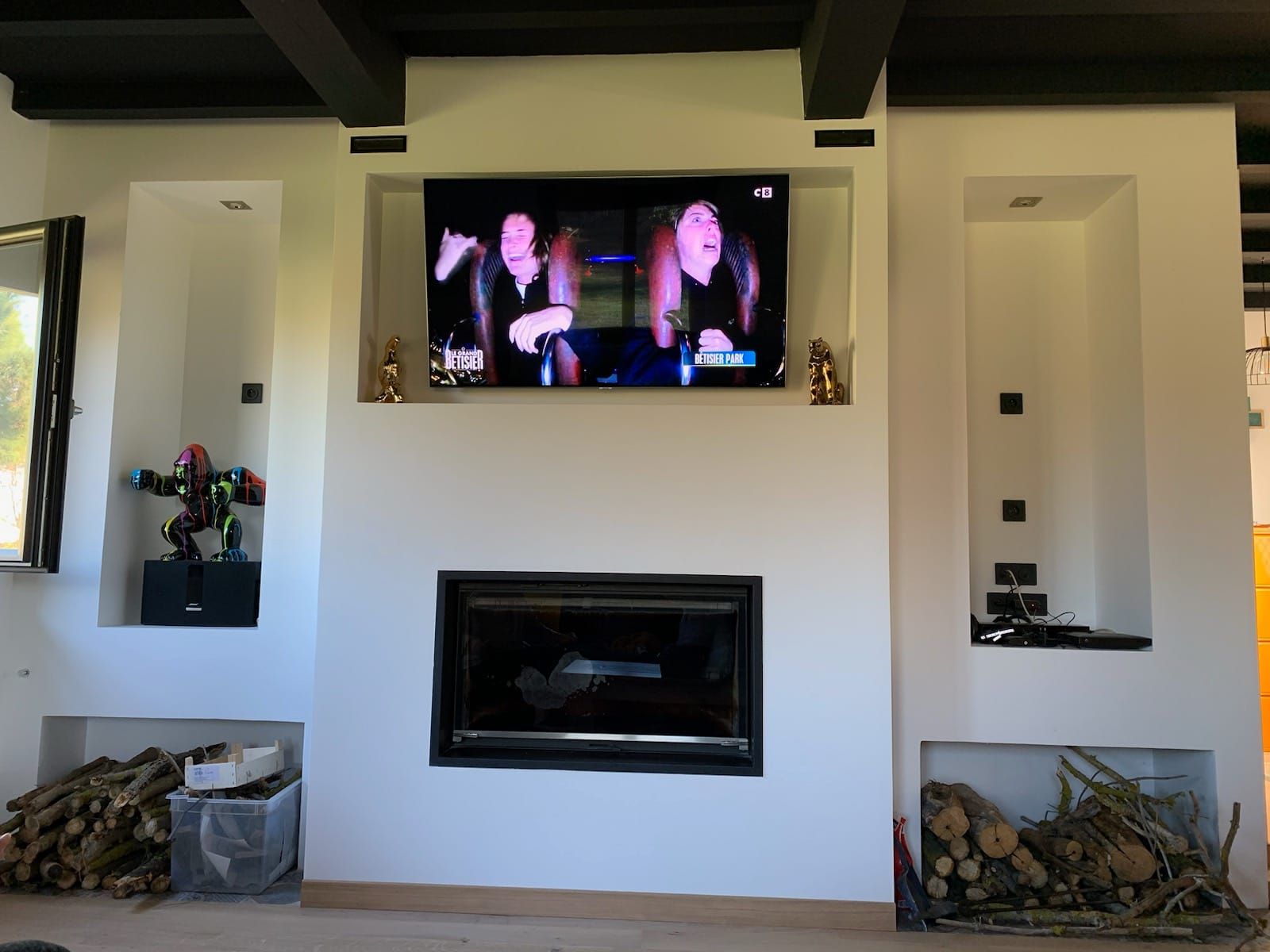 Cheminée blanche avec télévision intégrée et alcôves ; bois de chauffage empilé en dessous. Deux femmes à la télévision.