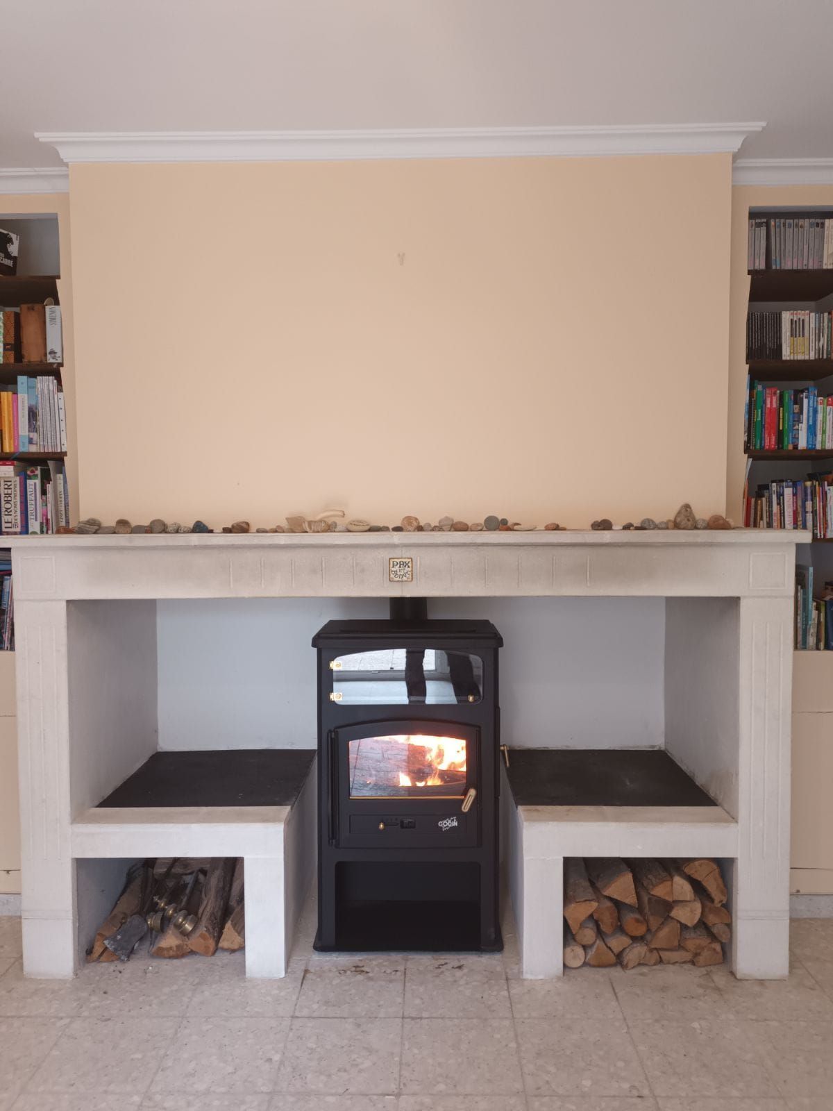 Cheminée avec poêle à bois allumé, rangement pour le bois de chauffage et bibliothèques encastrées contre un mur jaune pâle.