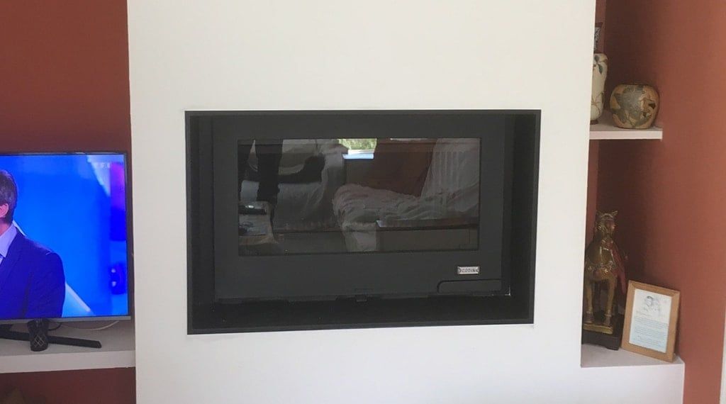 Cheminée encastrée dans un mur blanc. La vitre noire reflète la pièce. Étagères avec objets à droite et télévision à gauche.