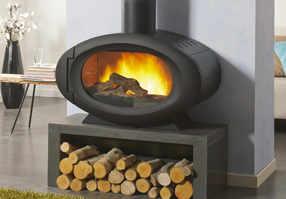 Cheminée ovale noire avec feu, bûches et socle de rangement en pierre.