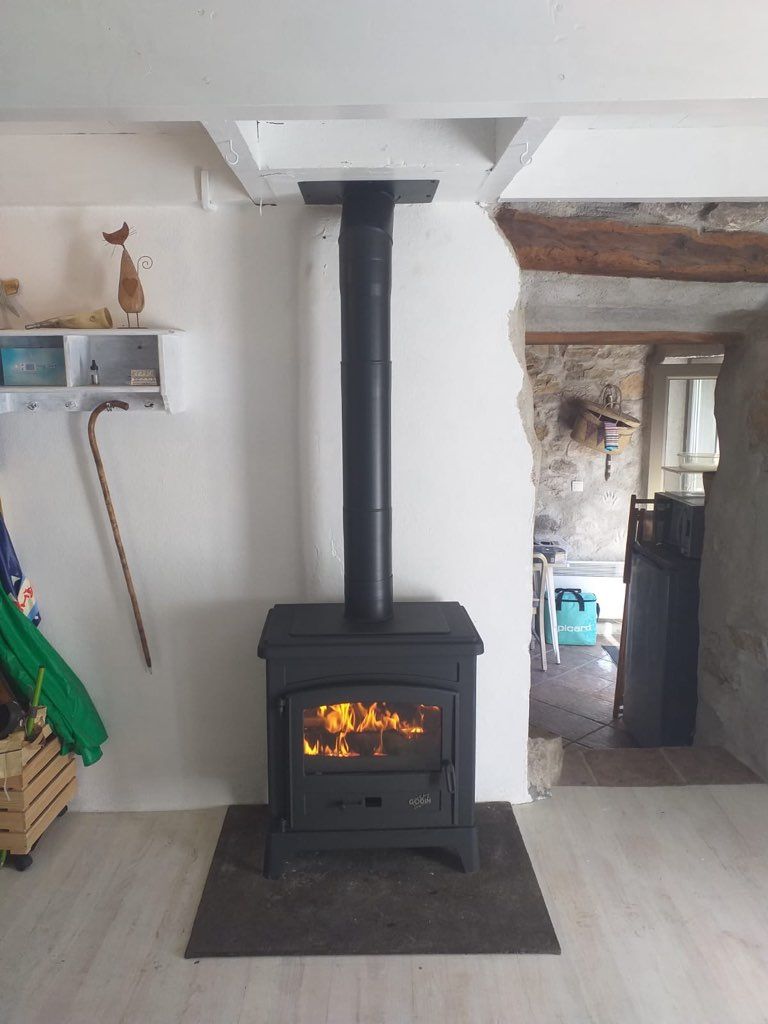 Un poêle à bois noir allumé, avec un conduit de cheminée, dans une pièce aux murs blancs et aux poutres apparentes.