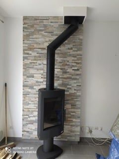 Poêle à bois noir avec conduit de fumée incliné, adossé à un mur à motifs de briques.