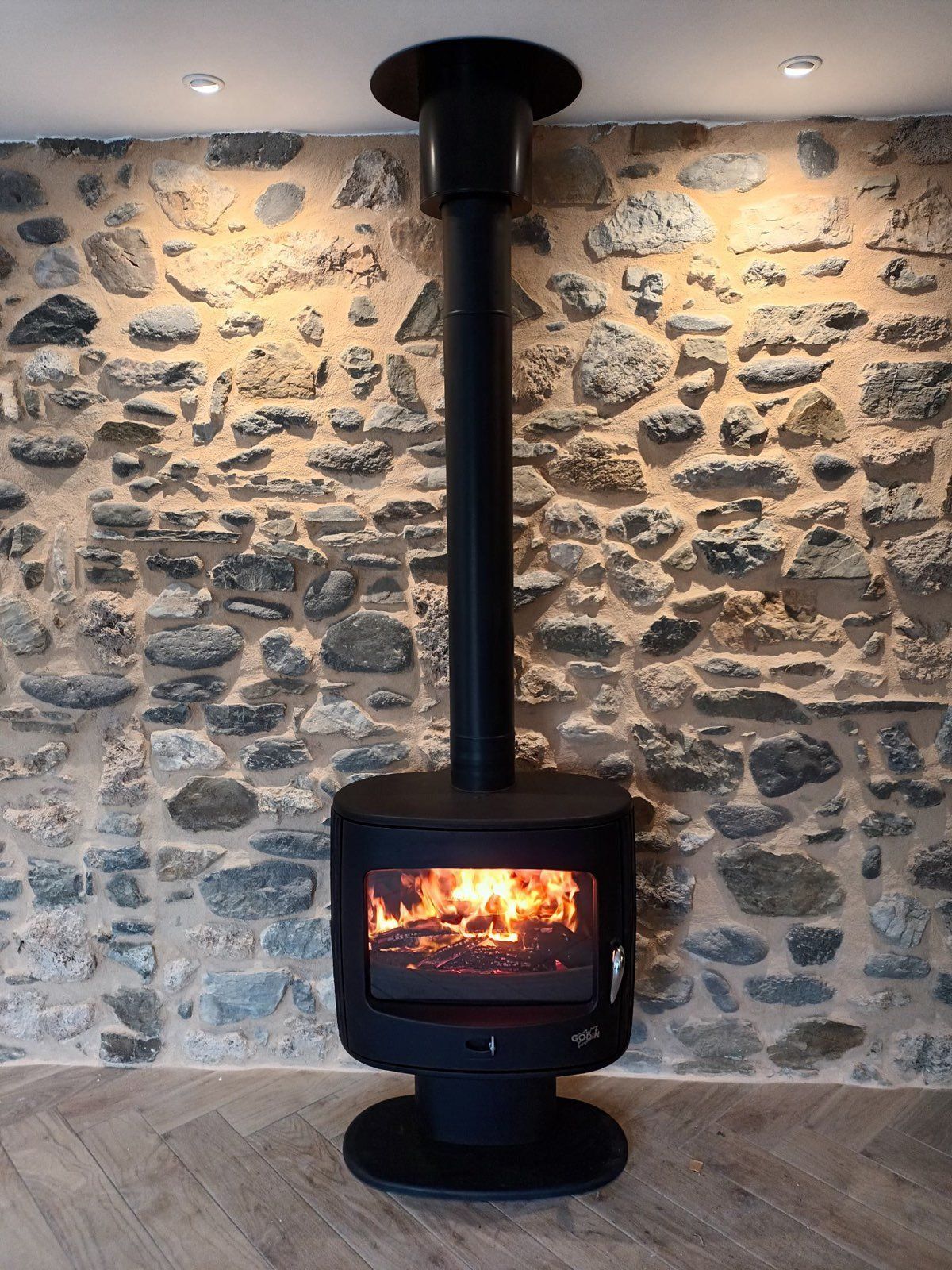 Poêle à bois noir avec feu allumé, adossé à un mur de pierre. La cheminée monte jusqu'au plafond.