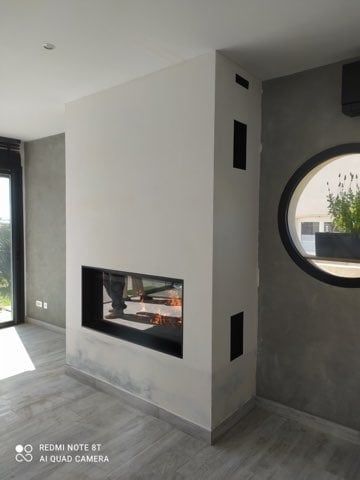 Cheminée moderne à flammes, encastrée dans un mur blanc avec une ouverture encadrée de noir. Une fenêtre ronde est visible.