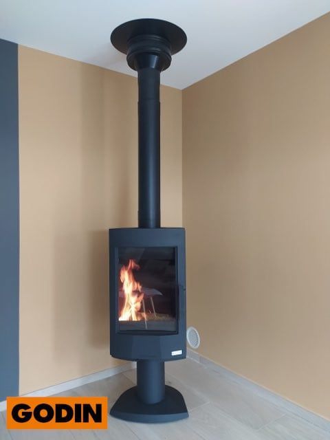 Poêle à bois Godin noir à flammes, conduit de fumée noir, installé dans un coin d'une pièce aux murs beiges et gris.