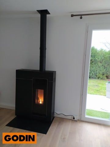 Poêle à granulés Godin noir avec flammes allumées, contre un mur blanc et une porte vitrée donnant sur un jardin.