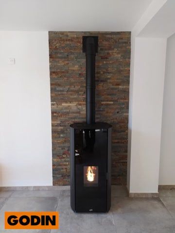 Poêle à granulés Godin noir avec un feu allumé, contre un mur de pierre, dans une pièce.