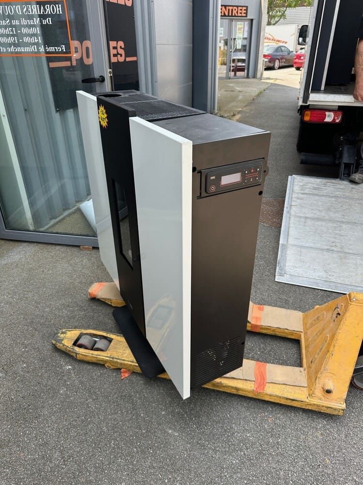 Poêle à granulés sur une palette à l'extérieur d'un bâtiment. Noir et blanc, panneau de commande visible.