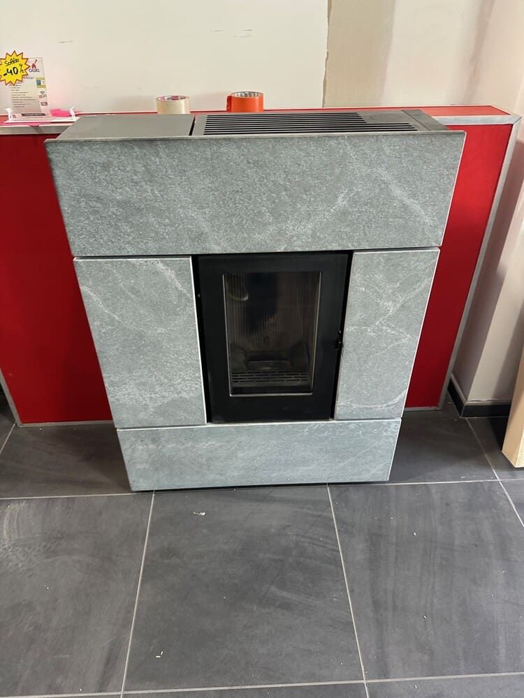 Poêle à granulés revêtu de pierre grise avec porte noire, posé sur un sol carrelé gris foncé. Mur rouge en arrière-plan.