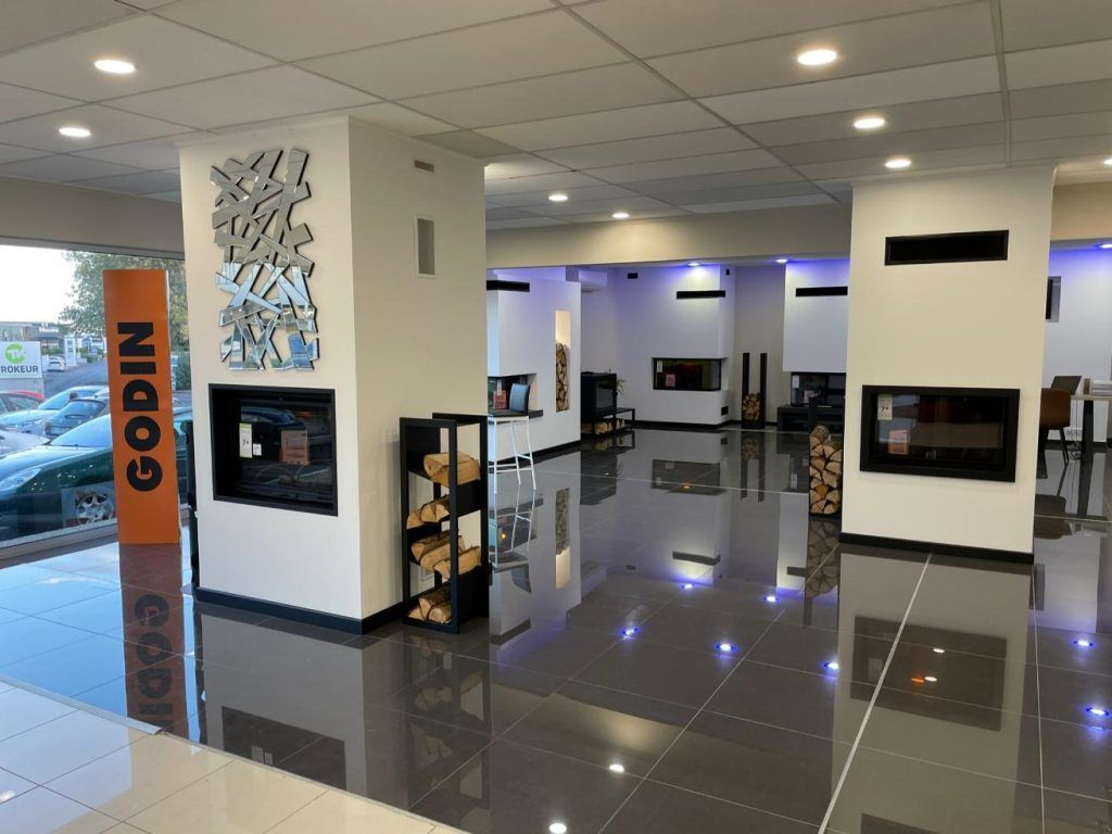 Intérieur d'une salle d'exposition présentant des cheminées modernes, un râtelier à bûches noir et une enseigne GODIN.