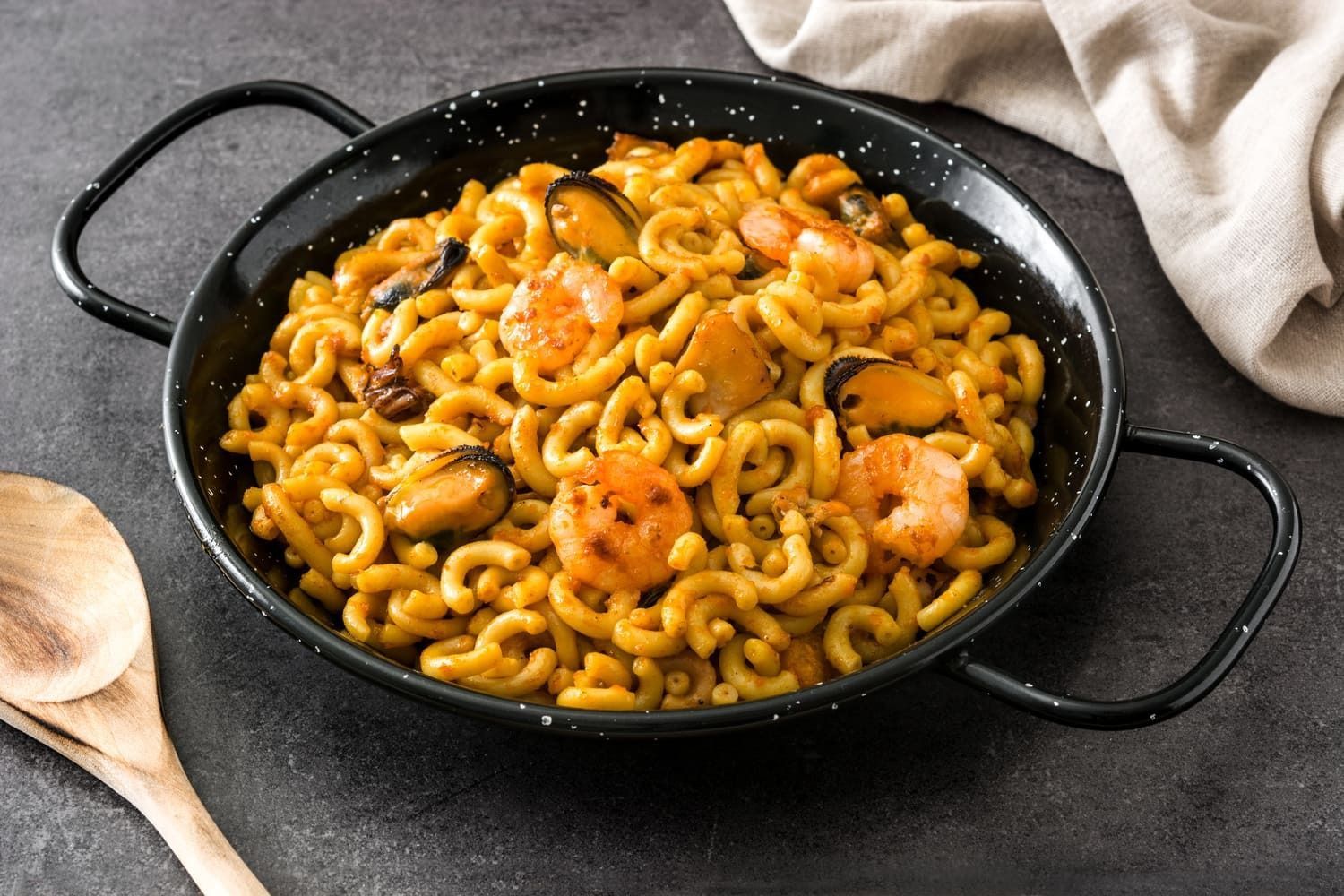 Una sartén llena de macarrones y camarones al lado de una cuchara de madera.