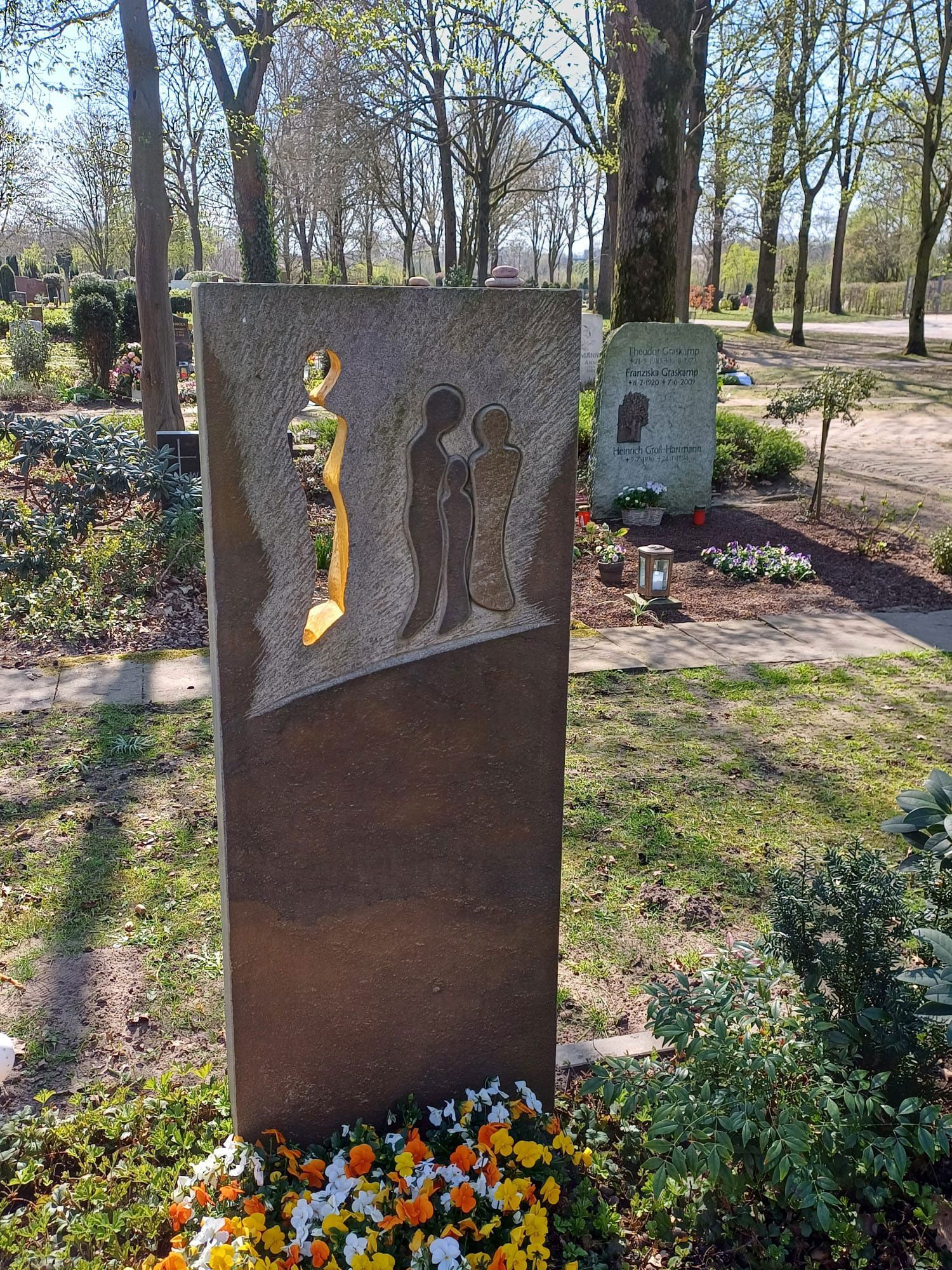 Ein Grabstein auf einem Friedhof mit Blumen und Bäumen im Hintergrund.