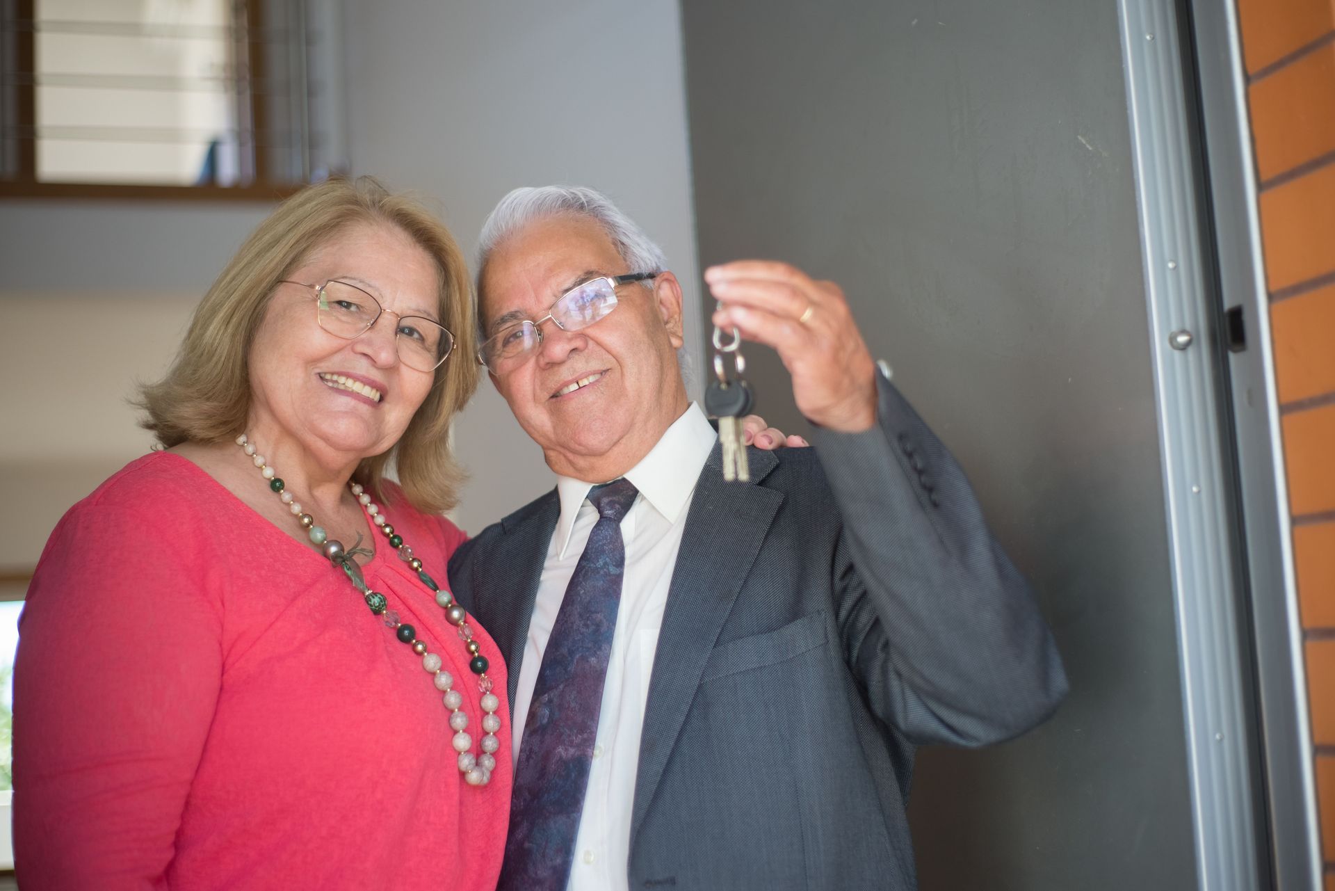 Una pareja sonriente está de pie en una puerta; el hombre sostiene unas llaves y la mujer lleva una blusa rosa.