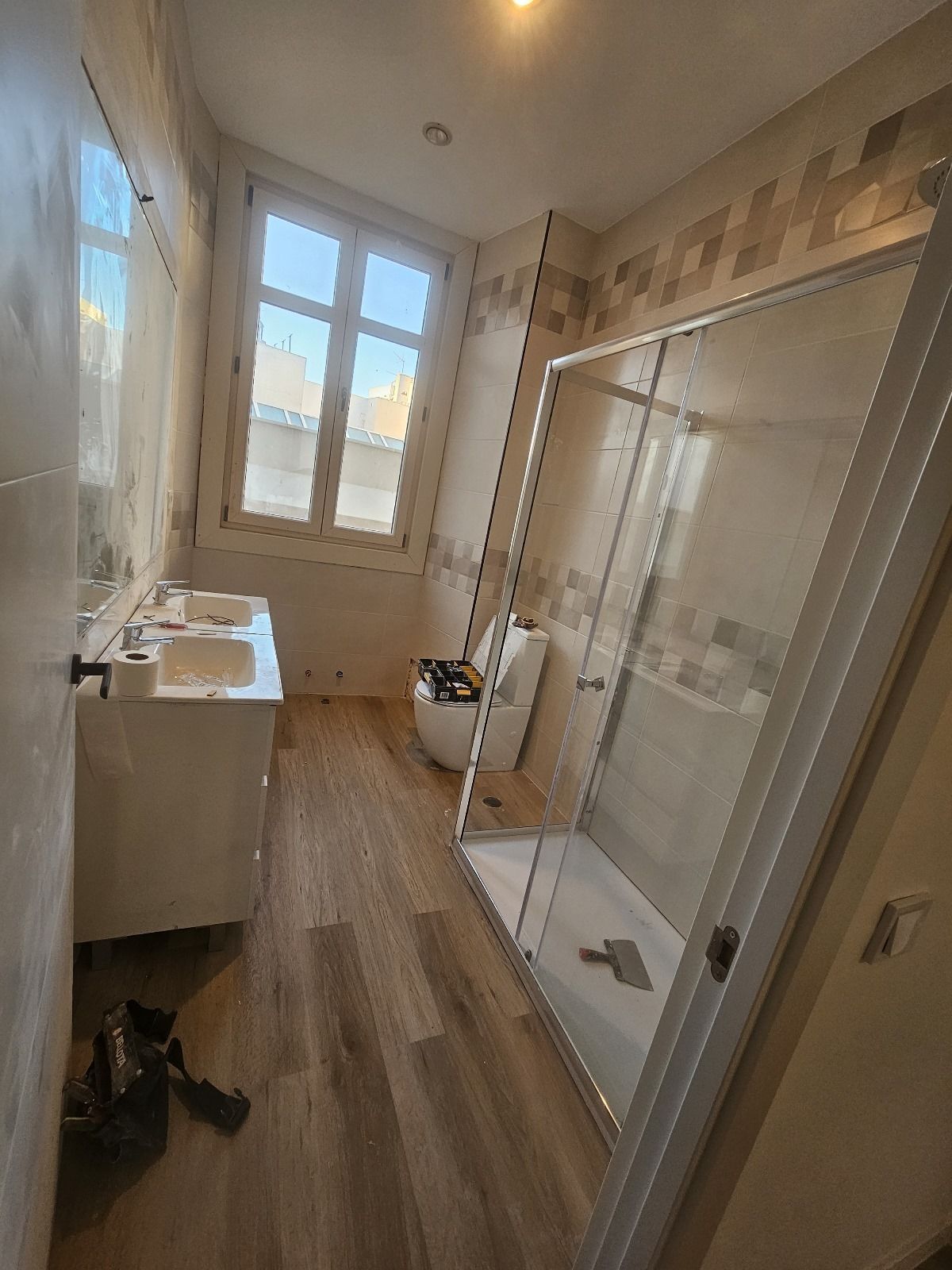 Baño con ducha, inodoro, lavabo y ventana; azulejos beige y suelo de madera.