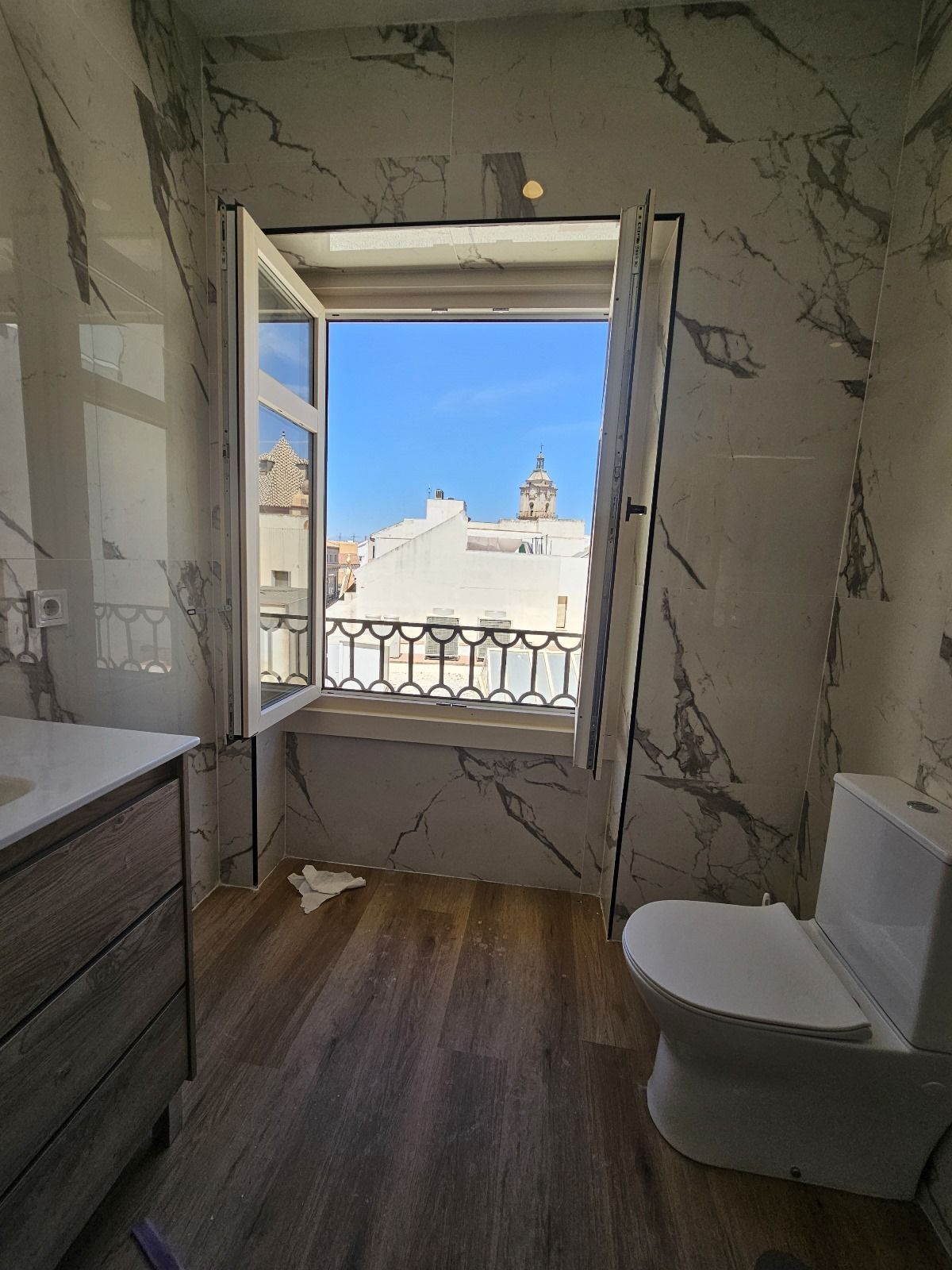 Baño con azulejos de mármol, ventana abierta y vista de edificios blancos bajo un cielo azul.