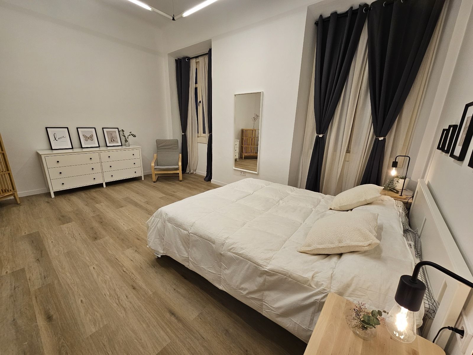 Dormitorio con cama, cómoda, espejo de cuerpo entero y cortinas. Suelo de madera clara. Paredes y ropa de cama blancas.