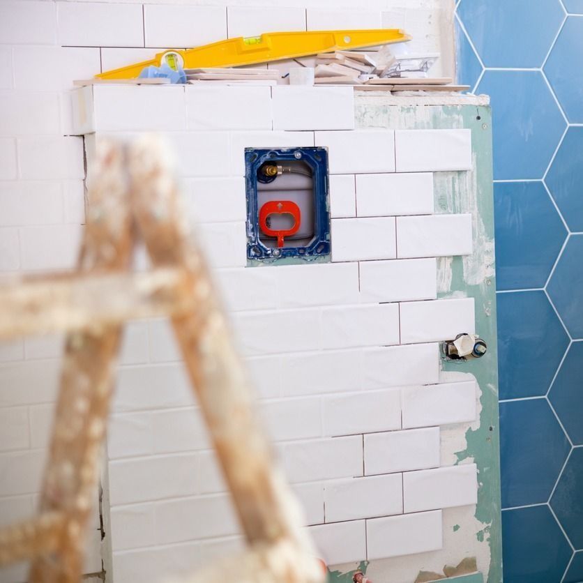 Pared de baño en construcción con azulejos blancos, una caja de enchufe azul y una escalera en primer plano.