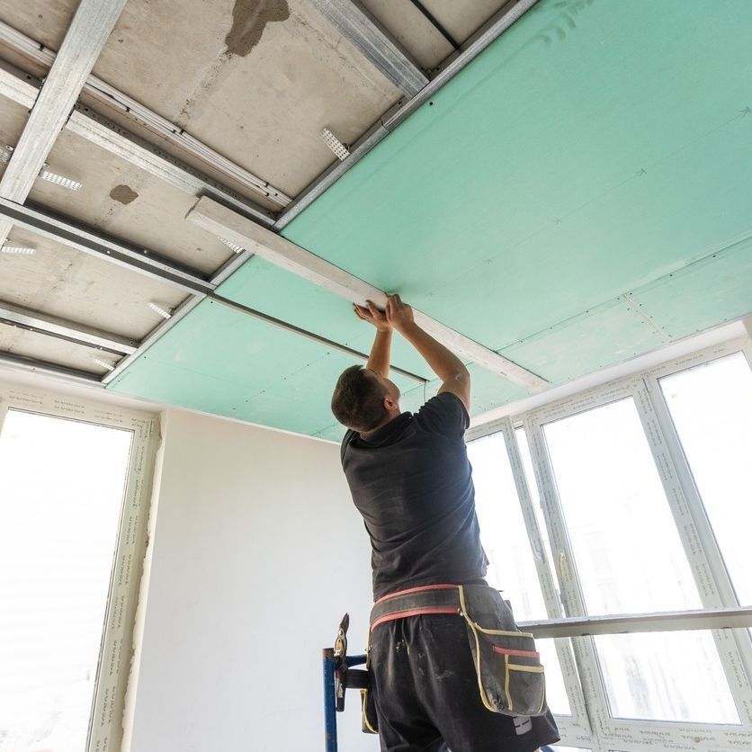 Un trabajador instalando paneles de yeso verdes en un techo con estructura de metal en una habitación con ventanas grandes.