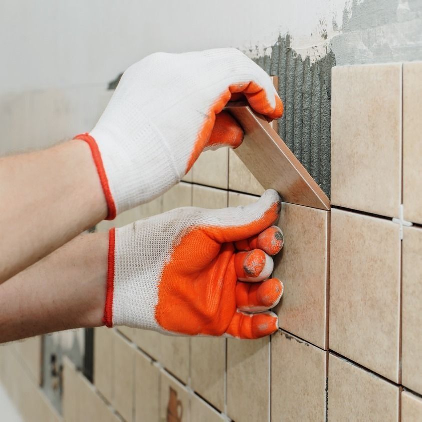 Manos con guantes de trabajo recubriendo una pared con azulejos de color beige.