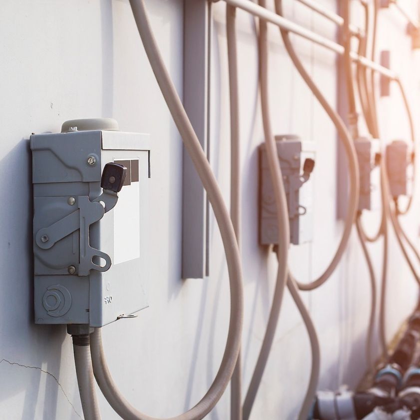 Cajas eléctricas y conductos grises sobre una pared blanca; la luz del sol proyecta un resplandor.