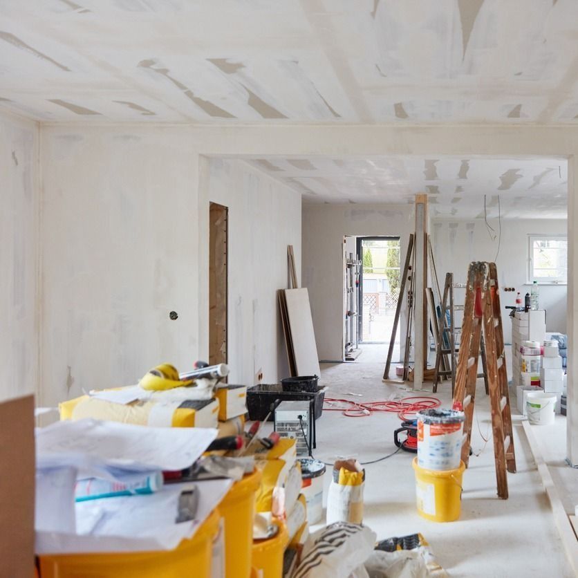 Interior de una casa en remodelación, con herramientas, materiales y escaleras. Paredes y techo blancos, puerta a otra habitación.