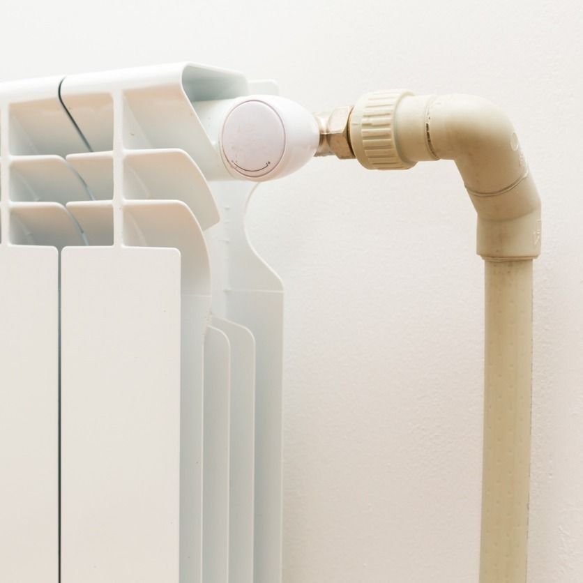 Radiador blanco con válvula y tubo beige contra una pared blanca.
