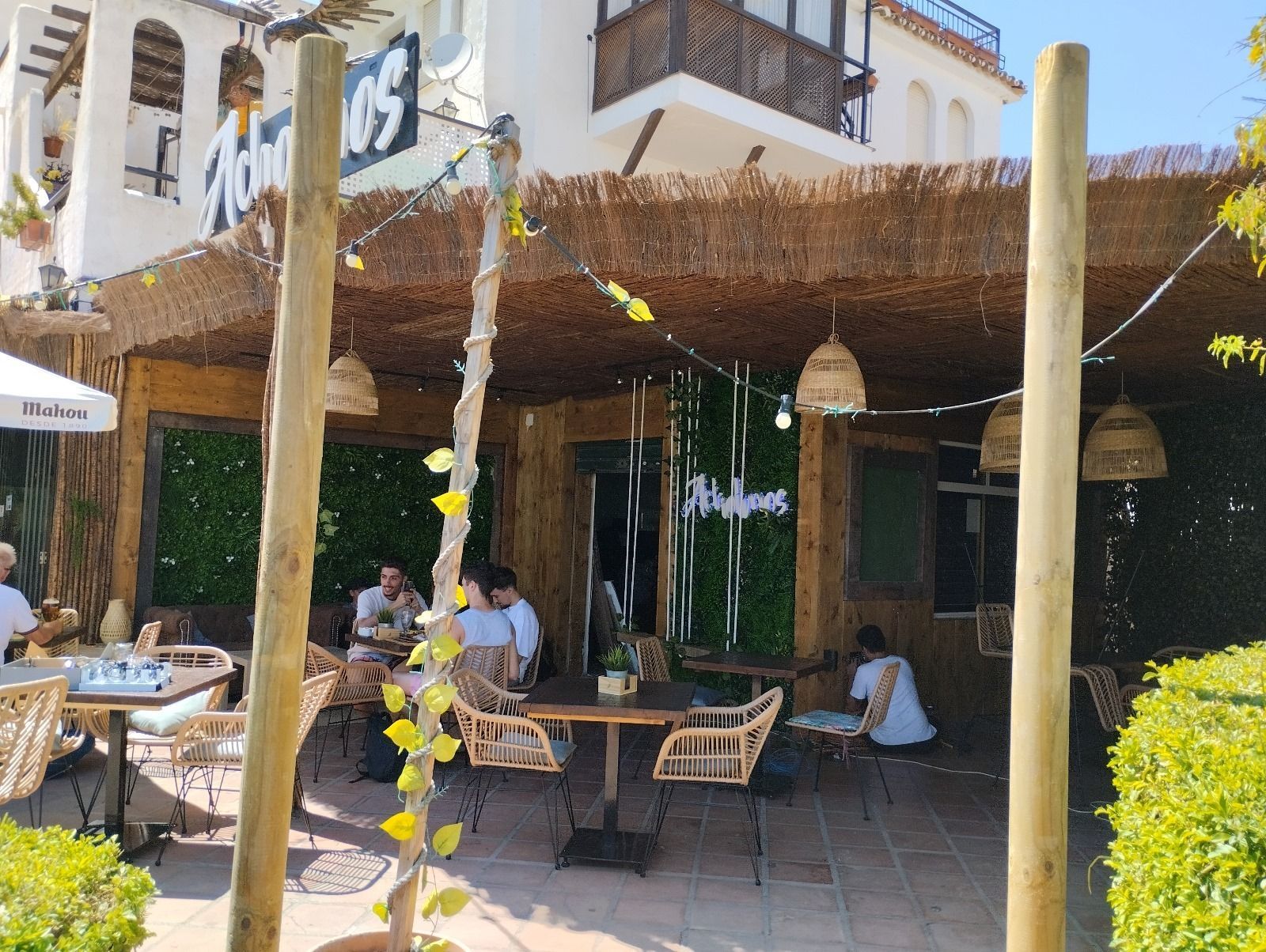 Cafetería al aire libre con techo de paja y lámparas colgantes. La gente cena en mesas; día soleado.