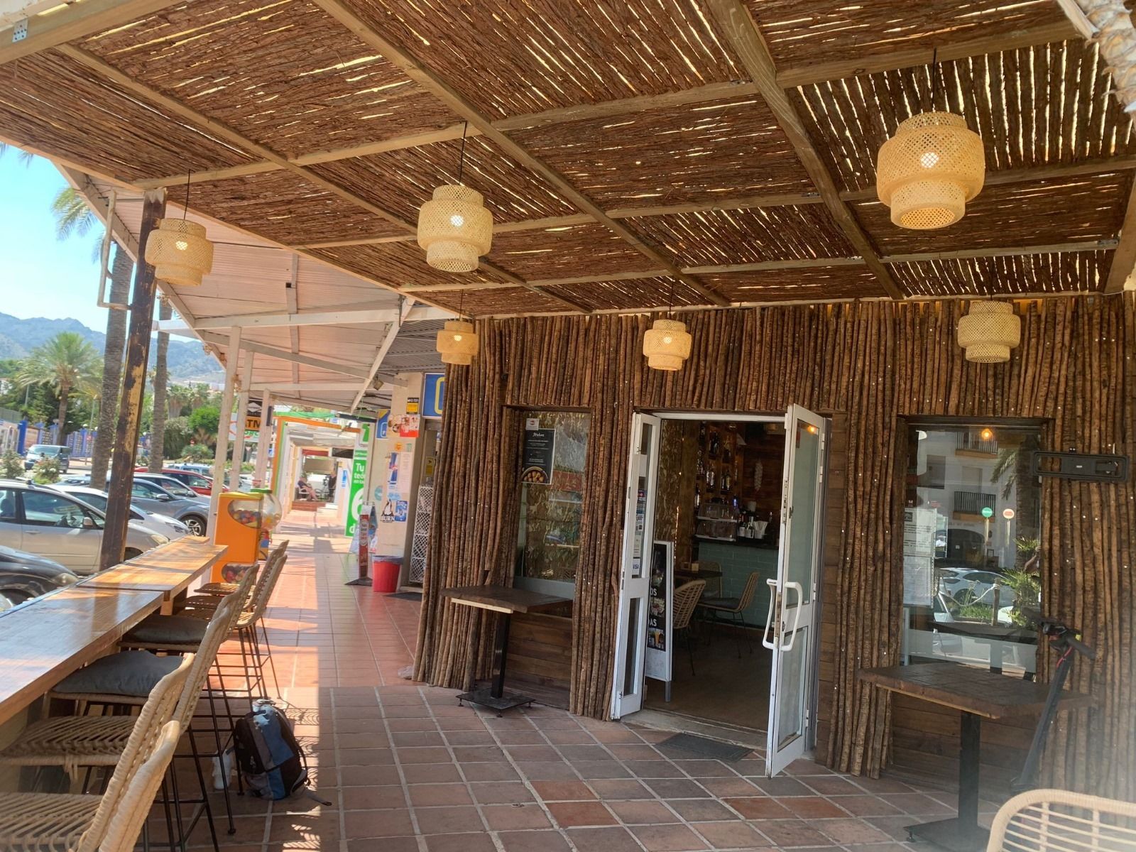 Exterior de cafetería con techo de paja, paredes de bambú, puertas abiertas, luces colgantes redondas y asientos en la acera.