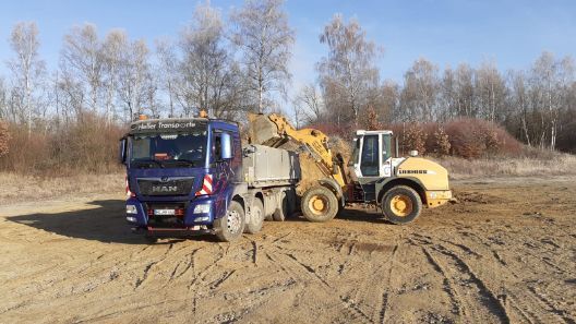 kleiner gelber Bagger schüttet etwas auf den Ladeanhänger eines Lkw