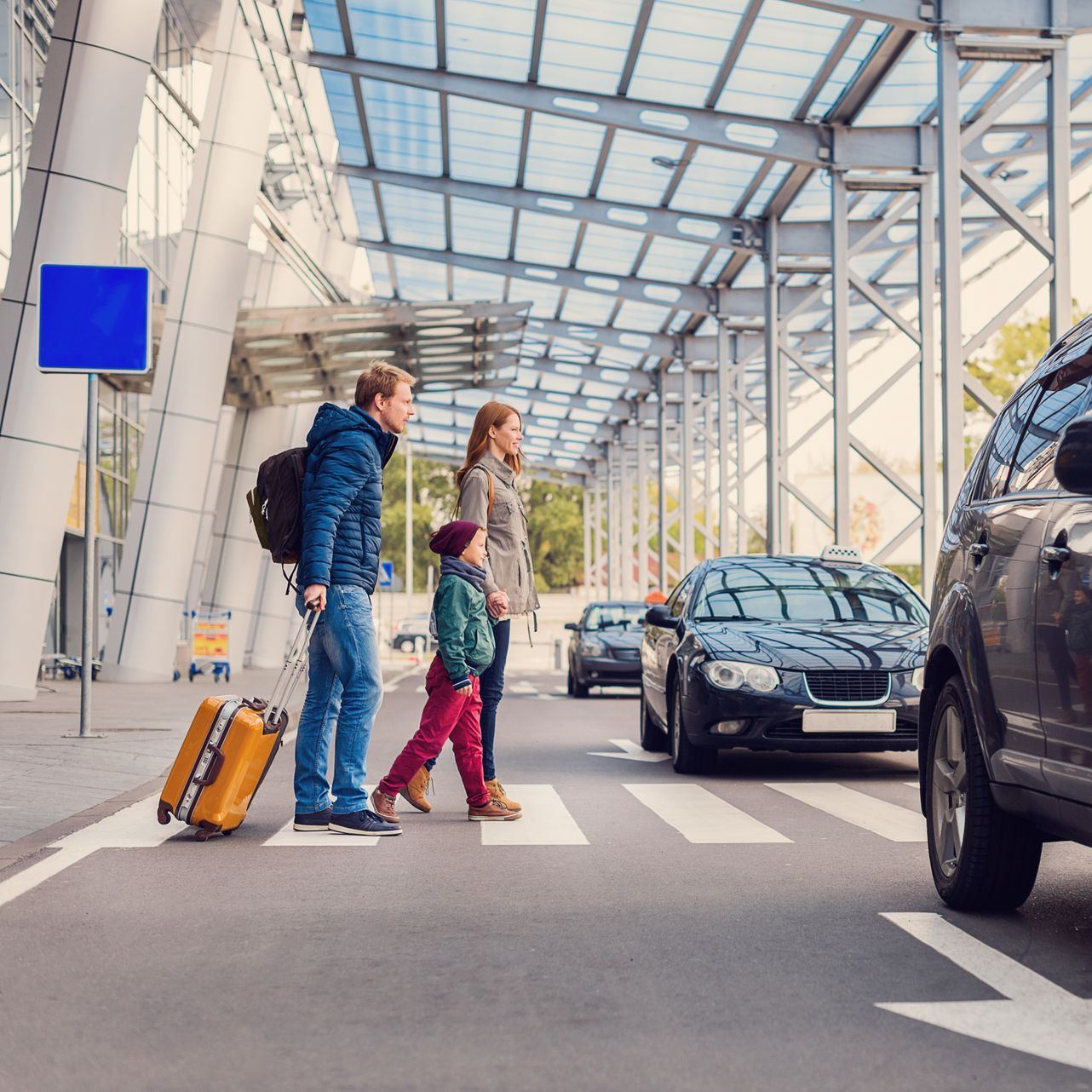 Une famille traverse un passage piéton à l'aéroport, des voitures se trouvent à proximité.