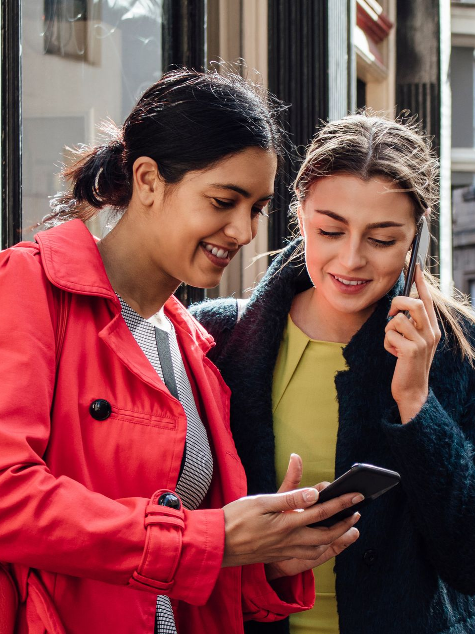Deux femmes regardent un téléphone en ville ; l'une tient le téléphone, l'autre parle au sien, toutes deux souriantes.