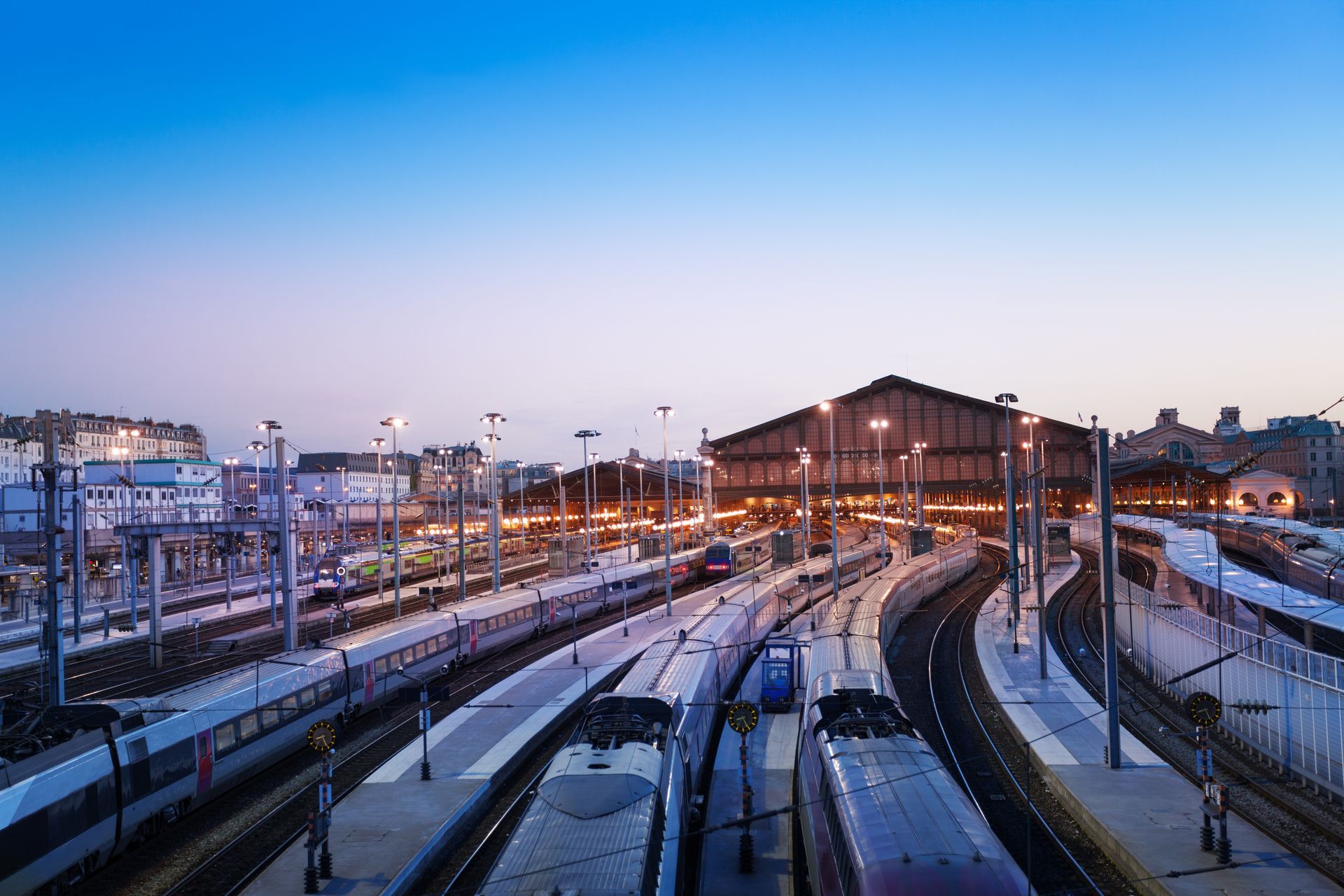 Au crépuscule, la gare voit converger plusieurs voies ferrées vers un grand bâtiment aux fenêtres éclairées.
