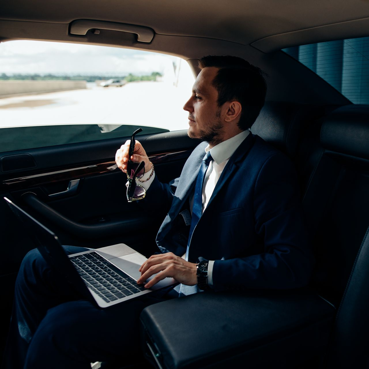 Un homme en costume, assis dans une voiture, regarde par la fenêtre, tient son téléphone et utilise un ordinateur portable.