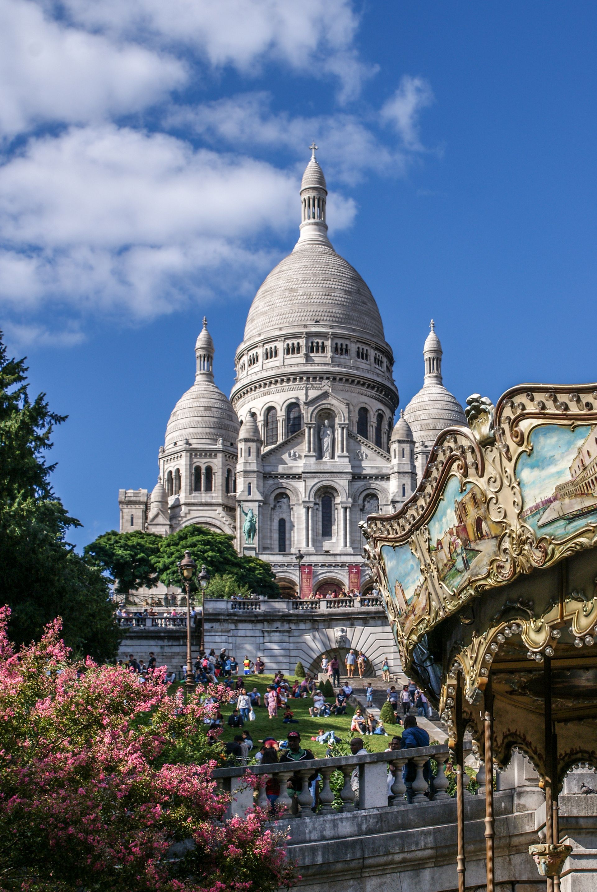 La basilique du Sacré-Cœur, à Paris, se dresse au-dessus d'un carrousel et de fleurs roses épanouies, sur un ciel bleu parsemé de nuages.
