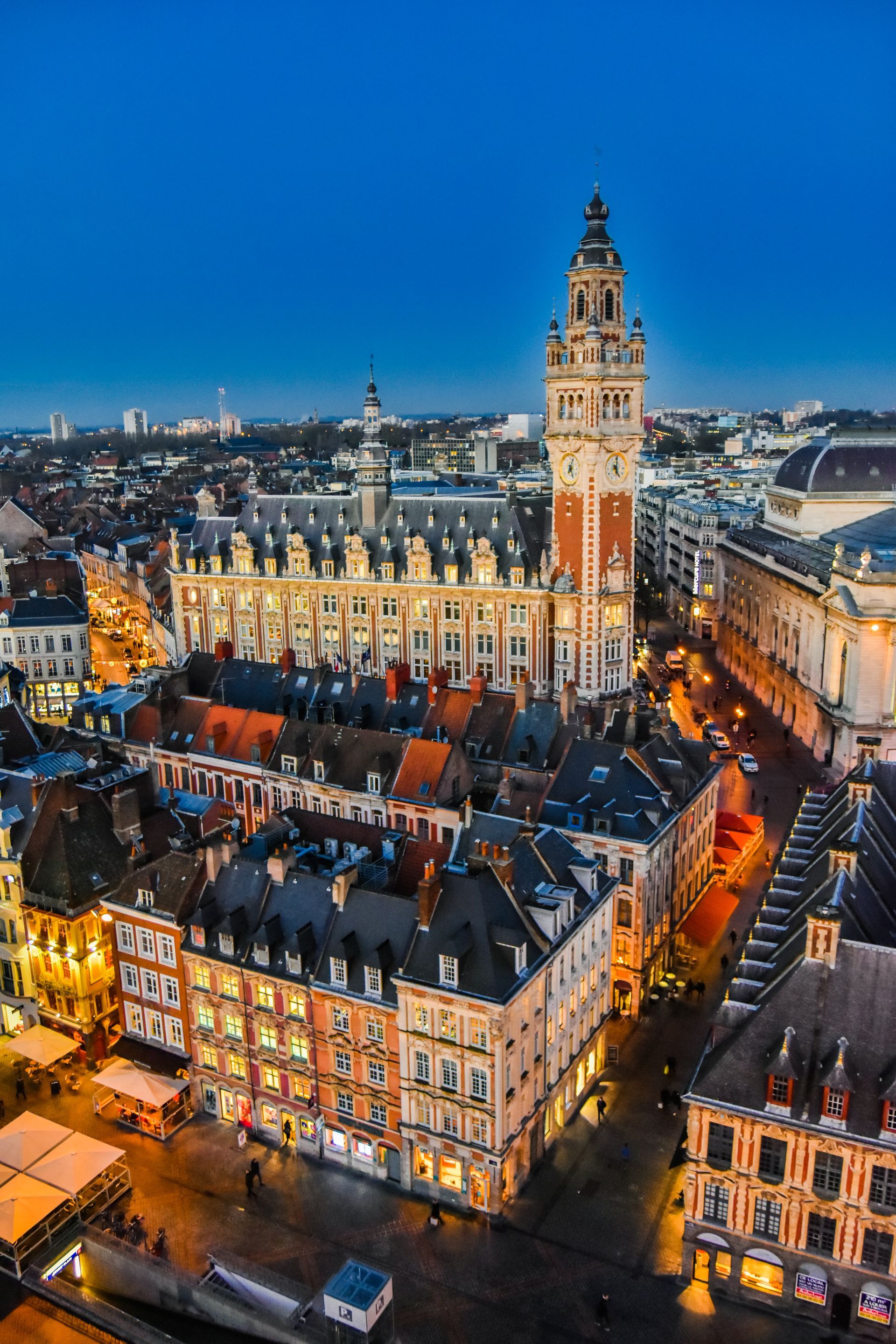 Vue de l'hôtel de ville de Lille, en France, au crépuscule. Les bâtiments aux toits orange et rouges sont illuminés.