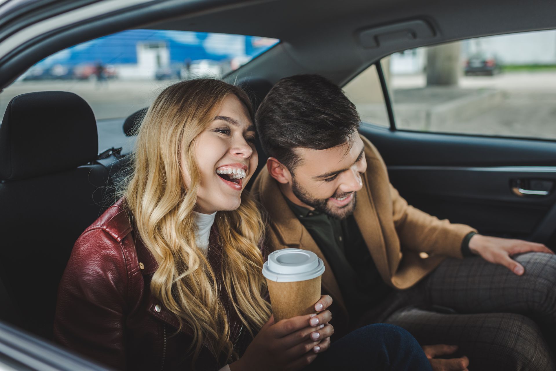Un couple rit dans une voiture, la femme tient un café, l'homme tend la main, tous deux sourient.