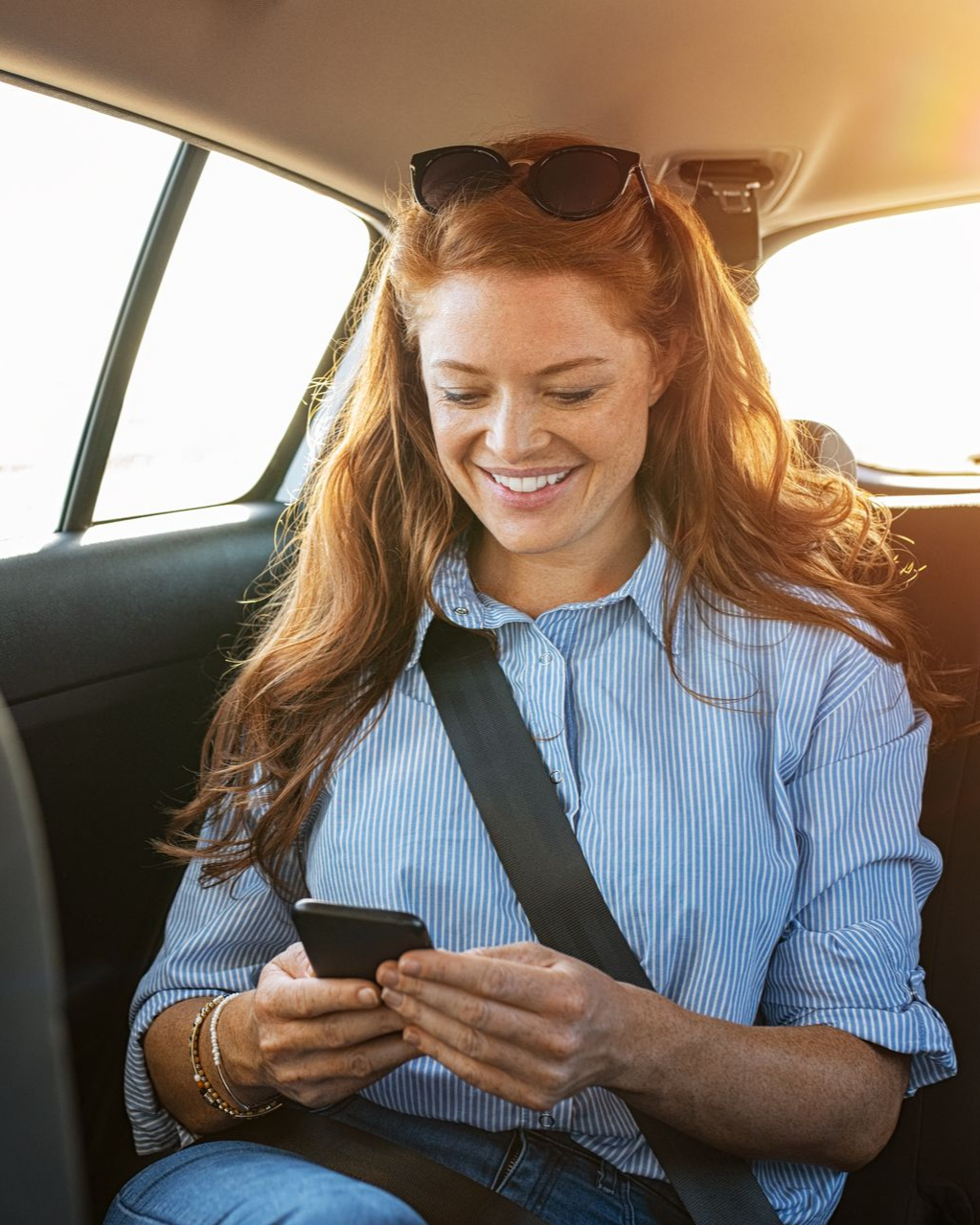 Une femme rousse sourit en utilisant son téléphone dans une voiture. Elle porte des lunettes de soleil et sa ceinture de sécurité.